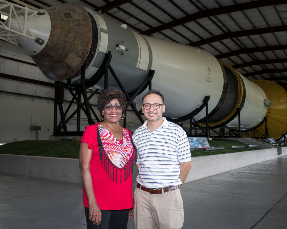 A Black woman and an Indian man stand in front of a Saturn V rocket that is laying on its side in an exhibit area.